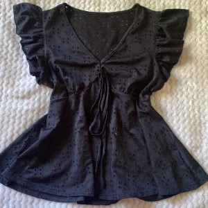 Svart blus med volangärm och knyt - Supersöt svart blus med v-ringning, volangärmar och knyt framtill. Tunt bomullstyg med broderade hål för en luftig känsla. Perfekt till jeans eller kjol för en trendig look. Endast använd ett fåtal gånger.