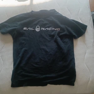 Svart Sail Racing t-shirt - Svart t-shirt från Sail Racing med vit logga tryckt över bröstet. Klassisk rund halsringning och korta ärmar. Tillverkad i mjuk bomull, perfekt för en avslappnad och sportig stil.