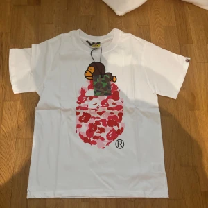 BAPE t-shirt - Vit t-shirt från BAPE med ikoniskt aptryck framtill. Stor grafisk apa med camo-detaljer och rosa-röda mönster på bröstet. Klassisk passform och crewneck. Perfekt för dig som gillar streetwear och vill sticka ut. Passar som M. ⚠️1:1⚠️helt oanvänd.