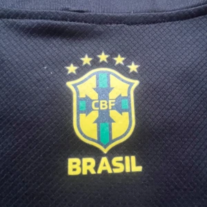 Brasil svart fotbollströja Nike - Snygg svart Brasilien fotbollströja från Nike med ikoniskt CBF-märke på bröstet och unikt konstnärligt tryck av Kristusstatyn och brasilianska motiv. Tröjan är kortärmad och tillverkad i lätt, ventilerande funktionsmaterial. Perfekt för dig som vill sticka ut på planen eller läktaren.