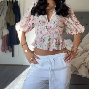Blommig croppad blus med puffärm - Superfin blommig blus i ljusa färger med v-ringning och croppad passform. Toppen har puffiga trekvartsärmar och smockad midja som ger en söt och feminin siluett. Perfekt för vår och sommar när du vill känna dig extra fräsch.