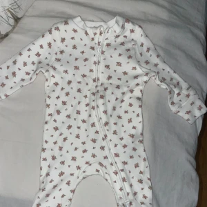 Vit blommig byxdress för baby - Supersöt vit byxdress med små blommor i rosa och rött. Långärmad och helkroppsmodell med dragkedja framtill som gör den enkel att ta på och av. Perfekt för mysiga dagar hemma eller som pyjamas.oanvända 