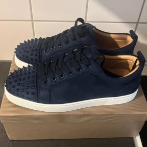 Helt nya! Loubotins med mocka. Pris kan diskuteras - Helt oanvända och Snygga mörkblå sneakers från Christian Louboutin i mocka med klassiska röda sulor. Skorna har rund tå, snörning och coola nitar framtill på tån för en edgy look. Insidan är ljusbeige och sulan är vit med röd undersida. Perfekta för dig som vill sticka ut. ( säljes pga fel storlek ) ( skriv ifall ni har några frågor eller funderingar! )
