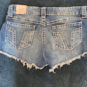 Lågmidjade jeansshorts från Gina Tricot - Snygga blå denimshorts från Gina Tricot med låg midja, slitna detaljer och fransig kant. Aldrig använt.