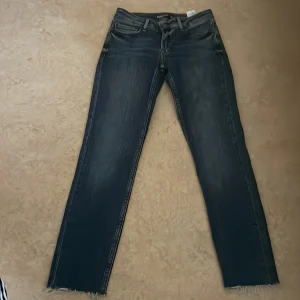 Superskinny blå jeans Sydnee 25 - Superskinny jeans i mörkblå tvätt från Sydnee, modell med smal passform och råa, fransiga benslut. Klassisk femficksdesign och dragkedjegylf. Perfekta för dig som gillar tajta jeans med lite edge.