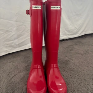 Röda Hunter stövlar i gummi - Snygga röda stövlar från Hunter i glansigt gummi med klassisk logga framtill och justerbart spänne på sidan. Perfekta för regniga dagar och festivaler. Hög modell              ❤️❤️