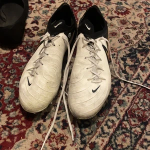 Nike Phantom vita fotbollsskor - Säljer ett par Nike Phantom fotbollsskor i vitt med svarta detaljer och guld på sidan. Skorna har snörning och en rund tå, samt syntetmaterial för bra grepp på planen. Perfekta för dig som vill sticka ut med stil på matchen.