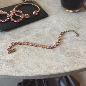 Caroline Svedbom armband - Snyggt armband i guldton med glittrande rosa stenar som fångar ljuset. Kedjan har små länkar och ett klassiskt lås, perfekt för att ge din look lite extra färg och bling. Stenarna är runda och sitter tätt längs hela armbandet.