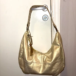 Guldig axelväska i skinn med tofs - Säljer en snygg axelväska i guldigt skinn med bred axelrem och coola nitar. Väskan har dragkedja upptill och en dekorativ tofs i guld. Perfekt storlek, mått 40*30  cm,  för allt du behöver och ger en lyxig touch till din outfit.