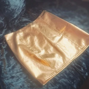 Guldig kortkjol i skinnlook - Supersnygg kortkjol i metallic guld med skinnkänsla. Kjolen har raka sömmar framtill och en clean, minimalistisk design som verkligen sticker ut. Perfekt för dig som vill ha en statement piece i garderoben.