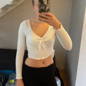 Vit croppad ribbad topp med knyt - Säljer en vit croppad topp med lång ärm och v-ringning. Toppen är ribbad och har en söt knytning framtill samt vågig kant nertill för en trendig look. Perfekt att styla med jeans eller kjol.🤍🤍
