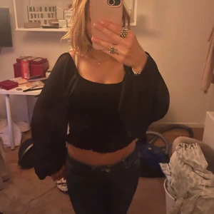 Svart croppad blus med puffärmar - Säljer en svart croppad blus med vida puffärmar och smockad resår vid midjan. Toppen har en fyrkantig ringning och är tillverkad i ett lätt, flowy tyg som ger en snygg siluett. Perfekt att styla med jeans eller kjol för en trendig look.