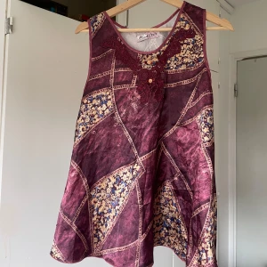 Vinröd blommig ärmlös linnetopp - Unik vinröd linnetopp med blommiga mönster i beige och blått. Toppen är ärmlös och har dekorativ spetsdetalj vid halsringningen. Lätt utsvängd passform och patchwork-inspirerat tyg som ger en cool och bohemisk vibe.