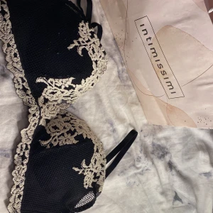 Svart spets-bh från Intimissimi - Supersnygg svart bh från Intimissimi med beige blommig spetsdetalj över kuporna och längs kanterna. Axelbanden är smala och bh:n har en elegant, feminin look med vackra broderade blommor. Perfekt för dig som gillar stilrena och romantiska underkläder.