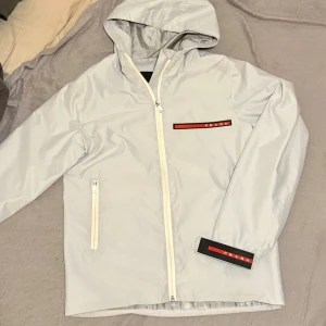 Ljusgrå vindjacka från Prada - Snygg ljusgrå vindjacka från Prada med huva och hel dragkedja framtill. Jackan har ikoniska röda Prada-loggor på bröstet och ärmen, samt en praktisk ficka med dragkedja. Tillverkad i glansig polyester som ger en clean och modern look. Size small, funkar för medium också 