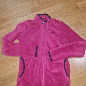Rosa fleecejacka från Rusta, stl S - Mysig rosa fleecejacka från Rusta med svarta detaljer vid krage, fickor och ärmslut. Jackan har hel dragkedja framtill och två fickor. Perfekt för kyliga dagar när du vill vara både bekväm och snygg.