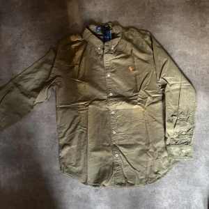 Olivgrön skjorta från Polo Ralph Lauren - Olivgrön skjorta från Polo Ralph Lauren med klassisk krage och knappar framtill. Skjortan har lång ärm och ikonisk orange broderad logga på bröstet. Tillverkad i mjuk bomull och passar perfekt till en avslappnad men ändå stilren look.