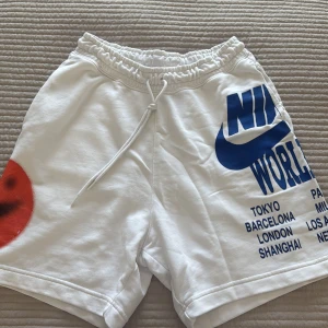 Vita Nike shorts med blå tryck - Snygga vita shorts från Nike med blått tryck och logga på ena benet samt röd detalj på andra sidan. Resår och snörning i midjan för skön passform. Perfekta för sommaren och träning eller chill.