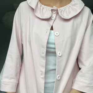 Ljusrosa kofta med volangkrage - Supersöt ljusrosa overshirt med stor volangkrage och vita knappar framtill. Jackan har trekvartsärmar På ryggen finns en diskret broderad detalj, (syns inte om man har utsläpp hår) 