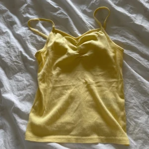 Gul ribbad linnetopp från H&M Divided - Säljer en ljusgul ribbad linnetopp från H&M Divided. Toppen har smala axelband, lätt rynk vid bysten och är tillverkad i mjuk bomull. Perfekt för varma dagar och enkel att styla med jeans eller kjol. Ej använd!