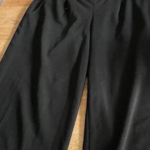 Svarta högmidjade wide byxor från JDY - Svarta byxor från JDY i storlek M/32. De har en loose och wide passform med raka, vida ben. Materialet känns mjukt och lätt, perfekt för en chill och stilren look. Passar dig som gillar bekväma och trendiga byxor. Noppighet på ena sidan av byxbenet.