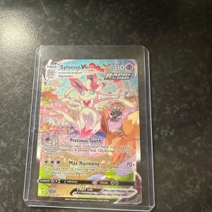 Sylveon VMAX - Cool och glittrig Pokémonkort med Sylveon VMAX! Perfekt för samlare och fans av Pokémon. Kortet har färgstark design och är skyddat i plastficka. Ett måste för dig som vill ha något unikt i din samling!