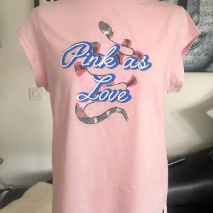 Rosa t-shirt med tryck från Coster - Snygg rosa t-shirt från Coster Copenhagen med coolt ormmotiv och texten 'Pink as Love' i blått framtill. T-shirten är i mjuk bomull, har korta ärmar och en relaxed passform. Perfekt för dig som gillar statement-plagg och unika prints.