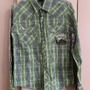 Grön rutig bomullsskjorta LAB Industries - Snygg grön rutig skjorta från LAB Industries i 100% bomull. Skjortan har långa ärmar, tryckknappar, två bröstfickor och coola tryckdetaljer både fram och bak. Perfekt för dig som gillar en avslappnad och trendig stil.