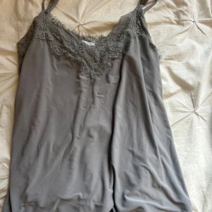 Grå linnetopp med spets från Amisu - Supersnygg grå linnetopp från Amisu med tunna axelband och v-ringning dekorerad med spetsdetaljer. Toppens material känns mjukt och svalt, perfekt för lager-på-lager eller som statement-piece. Spetsen ger en extra touch av elegans och trend.