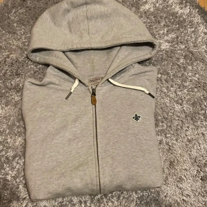 Grå hoodie från Morris med dragkedja - Snygg grå hoodie från Morris med dragkedja och vit snörning i huvan. Tröjan har en broderad logga på bröstet och klassisk passform. Perfekt för chill dagar och enkel att matcha med jeans eller joggers.
