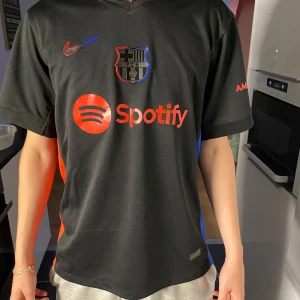 FC Barcelona tröja  - Snygg svart FC Barcelona matchtröja från Nike med Pedri och nummer 8 på ryggen. Tröjan har röda detaljer, Spotify-logga på bröstet och UNHCR-tryck bak. Tillverkad i lätt och ventilerande material, perfekt för fotbollsälskare.