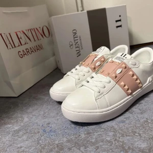 Valentino Garavani Rockstud sneakers - Snygga vita Valentino Garavani Rockstud sneakers i skinn med breda ljusrosa band och ikoniska nitar över vristen. Klassisk rund tå, platt sula och coola detaljer även på sulan. Perfekta för dig som vill sticka ut med exklusiv streetstyle.