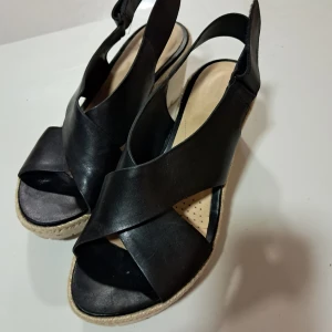 Svarta sandaler med kilklack från Clarks - Snygga svarta sandaler från Clarks i skinn med korsade remmar och öppen tå. De har en beige kilklack med flätad detalj och vit sula. Perfekta för sommaren och ger en stilren look till alla outfits.