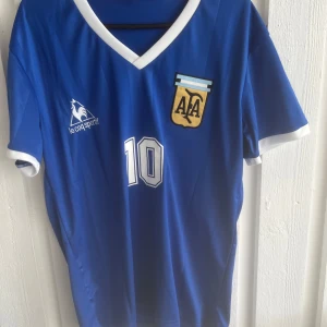 Blå Maradona Argentina tröja Le Coq Sportif - Snygg blå Argentina fotbollströja från Le Coq Sportif med nummer 10 och Maradona på ryggen. Tröjan har vita detaljer vid krage och ärmslut, samt AFA-emblem på bröstet. Perfekt för dig som älskar klassisk fotbollsstil.
