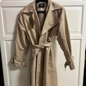 Beige trenchcoat från Bik Bok, S - Stilren beige trenchcoat från Bik Bok med klassisk krage, bälte i midjan och spännen vid ärmsluten. Jackan har två fickor framtill och är tillverkad i ett mjukt bomullsmaterial. Perfekt för lager-på-lager och snygg till alla outfits.