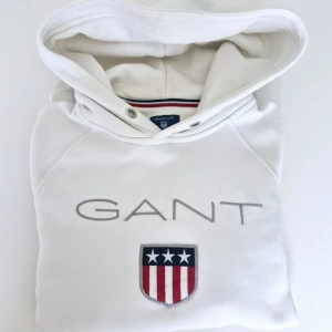 Vit hoodie från GANT med logga - Snygg vit hoodie från GANT med stor broderad logga och sköld på bröstet. Klassisk huva med snörning och känguruficka framtill. Perfekt för dig som gillar en clean och sportig stil. Tillverkad i mjuk bomull för maximal komfort.