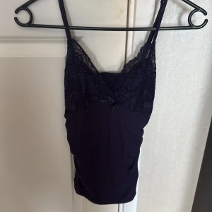 Mörkblå spetslinnetopp med rosett - Supersöt mörkblå linne-topp med smala axelband och v-ringning. Överdelen är dekorerad med blommig spets och en liten rosett framtill. Perfekt för lager-på-lager eller som statement-piece. Mjuk och stretchig känsla.