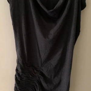 Svart kortklänning från Sirup - Svart kortklänning från Sirup med snygg drapering på ena sidan och lös passform. Klänningen har en enkel design med mjukt tyg och lätt fall. Perfekt för dig som gillar stilrena plagg med en twist.