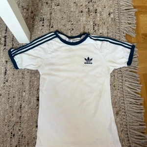 Vit Adidas t-shirt med mörkblå ränder - Vit t-shirt från Adidas med klassiska blå ränder längs axlarna och blå logga på bröstet. T-shirten har rund halsringning och blå kant vid ärmslut och hals. Perfekt för en sportig och avslappnad stil.