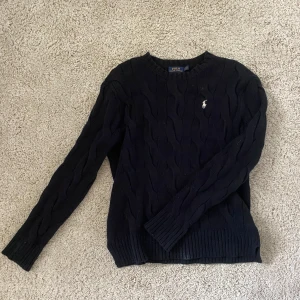 Svart kabelstickad tröja Polo Ralph Lauren - Kabelstickad svart tröja från Polo Ralph Lauren med klassisk rund halsringning och broderad logga på bröstet. Tröjan är långärmad och har ribbade muddar vid ärmslut och nederkant. Perfekt för dig som gillar stilren och tidlös design.