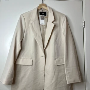 Beige kavaj från Bik Bok XS/S - Stilren beige kavaj från Bik Bok i rak modell med klassisk krage och knäppning framtill. Kavajen har fickor med lock och knappar vid ärmslut. 