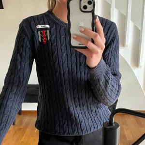Mörkblå kabelstickad tröja med broderad logga - Kabelstickad mörkblå tröja med rund hals och lång ärm. Tröjan har en broderad logga på vänster bröst och ribbade muddar vid ärmslut och nederkant. 