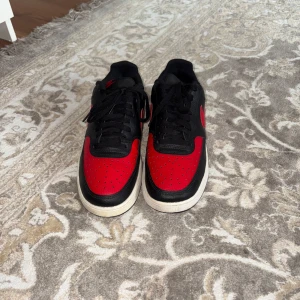 Nike Court vision sneakers  - Snygga Nike sneakers i svart och rött med klassisk vit sula. Skorna har lågt skaft, svart snörning och röd swoosh på sidan. Ovandelen är i skinn med perforerade detaljer på tån för extra stilpoäng. Perfekta för dig som gillar streetwear och vill sticka ut.