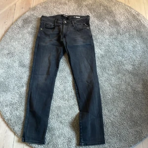 Replay Anbass svarta jeans, stl 32 - Säljer ett par svarta Replay Anbass jeans i slim fit. Jeansen har klassisk femficksdesign, dragkedjegylf och snygga detaljer på bakfickorna. Modellen är smal och sitter tajt längs benen. Perfekta svarta jeans för dig som gillar stilren look.