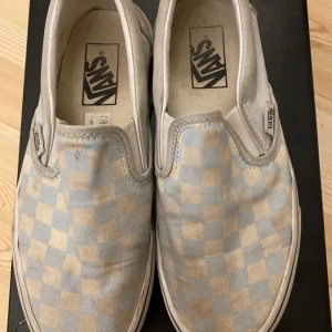 Vans storlek 37 - Klassiska Vans slip-on sneakers med rutigt mönster i ljusblått och vitt. Skorna har en låg profil, platt sula och är tillverkade i canvas. Perfekta för en avslappnad streetstil och lätta att bara hoppa i.