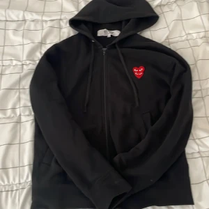 Svart hoodie Comme des Garçons Play - Svart hoodie från Comme des Garçons Play med dragkedja och huva. På bröstet sitter det ikoniska röda hjärtat med ögon. Tröjan har långa ärmar, fickor framtill och är tillverkad i mjuk bomull. Perfekt för dig som gillar streetwear och vill sticka ut.