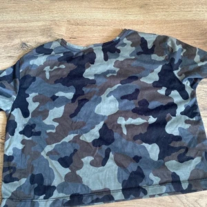 Camouflage t-shirt från Adidas - Säljer en croppad t-shirt från Adidas med klassiskt stort vitt tryck framtill. T-shirten har korta ärmar och coolt camouflage-mönster i grönt, brunt och svart. Perfekt för dig som gillar streetstyle och sportiga vibes.