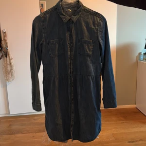 Mörk jeansklänning från GapKids - Snygg mörkblå jeansklänning från GapKids med klassisk skjortkrage, två bröstfickor och långa ärmar. Klänningen har knäppning framtill och är tillverkad i mjukt jeansmaterial. Perfekt för dig som gillar en avslappnad men cool stil.