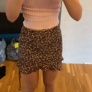 Brun blommig kjol - Supersöt brun kortkjol med vita blommor. Kjolen har snörning i sidan där man kan skjutstera längden i sidan. Den är gjord i ett lätt, flowy material som är perfekt för varma dagar.