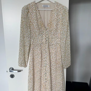 Blommig långklänning från Adoore - Supersöt långklänning från Adoore med små gula blommor på vit botten. Klänningen har v-ringning, långa ärmar och knappar framtill. Använd fåtal gånger, som nyskick! 